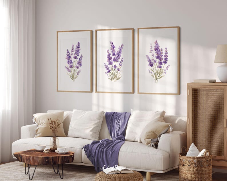 May include: Three framed art prints featuring watercolor paintings of lavender sprigs. The lavender is depicted in shades of purple and green against a white background. The frames are a light brown color. The prints are displayed above a white sofa.