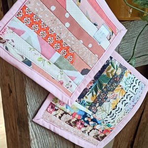 Könnte beinhalten: Zwei handgefertigte, gesteppte Topflappen mit Patchwork-Design. Die Topflappen weisen eine Vielzahl von Stoffen in Rosa-, Pfirsich- und Weißtönen mit Blumen- und Punktmustern auf. Jeder Topflappen ist mit einem rosa Rand versehen.