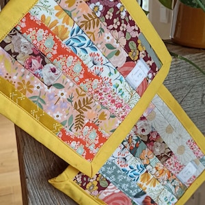 Könnte beinhalten: Zwei handgefertigte, quadratische, gesteppte Topflappen mit einem floralen Patchwork-Design. Der Patchwork-Stoff weist verschiedene Blumenmuster in Rosa-, Orange-, Blau- und Burgundertönen auf. Jeder Topflappen hat einen leuchtend gelben Rand und eine weiße Aufhängeschlaufe.