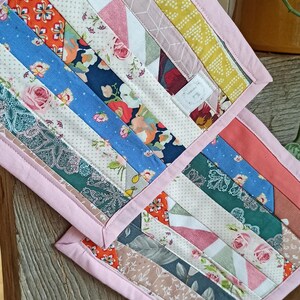 Könnte beinhalten: Zwei handgefertigte, gesteppte Topflappen mit rosa Rand. Die Topflappen haben ein Patchwork-Design mit verschiedenen Stoffmustern, darunter Blumen-, geometrische und unifarbene Muster. Die Stoffe umfassen Blau-, Rosa-, Gelb- und Grüntöne.