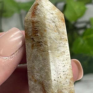 May include: A light tan crystal obelisk with reddish-brown and white veining. The crystal is held in a person's hand. The obelisk has a pointed top and a rectangular base. The background is blurred with green foliage.