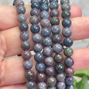 May include: A close-up of several strands of gemstone bead bracelets. The beads are a mix of colors, including gray, blue, green, and red. The beads are round and appear to be made of natural stone. The bracelets are held in a hand.