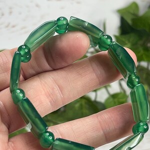 May include: A green beaded bracelet featuring cylindrical and spherical beads. The bracelet is made of polished, translucent green stones. The beads are strung together to form a circular shape. The bracelet is shown against a blurred background.