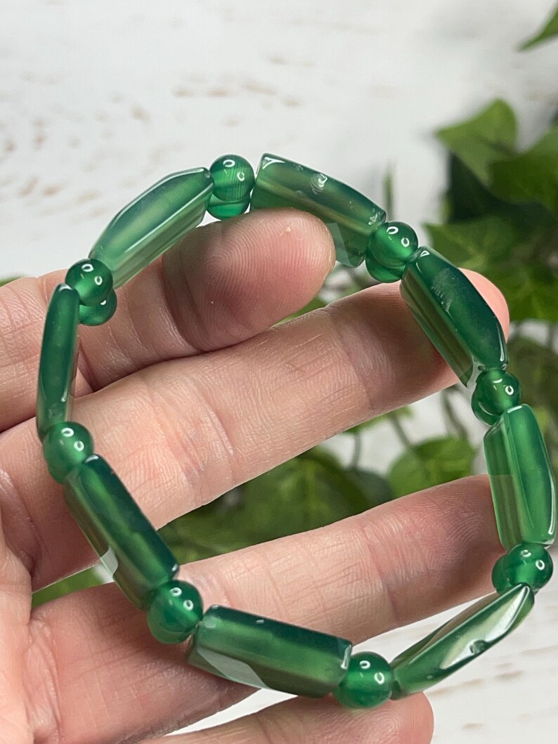 May include: A green beaded bracelet featuring cylindrical and spherical beads. The bracelet is made of polished, translucent green stones. The beads are strung together to form a circular shape. The bracelet is shown against a blurred background.