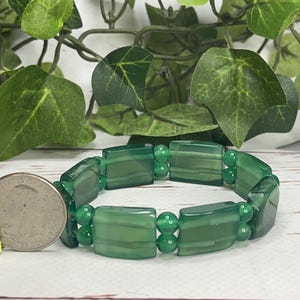 May include: A green beaded bracelet with rectangular and round beads. The bracelet is shown next to a quarter coin for size reference. The background includes green leaves and a white wooden surface.