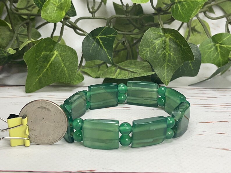 May include: A green beaded bracelet with rectangular and round beads. The bracelet is shown next to a quarter coin for size reference. The background includes green leaves and a white wooden surface.