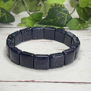 May include: A dark blue bracelet made of rectangular, polished stones with sparkling inclusions. The bracelet is displayed on a white wooden surface, with green foliage in the background. The bracelet is a jewelry item.