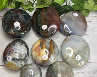 May include: Seven polished gemstones in various colors, including red, green, brown, gray, and yellow. Each stone has a unique pattern and is labeled with a letter from A to H. The stones are displayed on a white wooden surface.