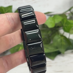 May include: A black rectangular link bracelet. The bracelet is made of glossy, polished black material. The links are connected to form a flexible band. The background features green foliage and a light-colored surface.