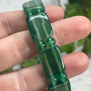 May include: A green beaded bracelet featuring rectangular and round beads. The rectangular beads have a translucent appearance with visible internal patterns. The bracelet is held up against a blurred background, highlighting the jewelry's color and design.