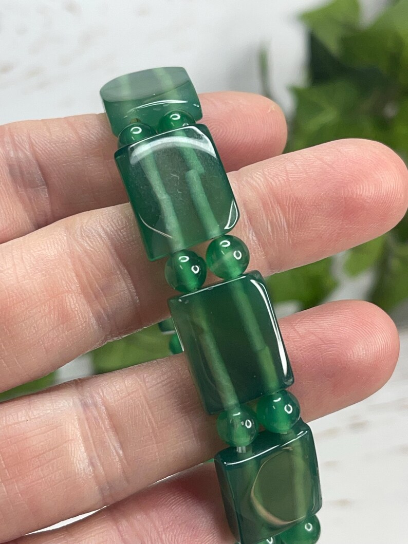 May include: A green beaded bracelet featuring rectangular and round beads. The rectangular beads have a translucent appearance with visible internal patterns. The bracelet is held up against a blurred background, highlighting the jewelry's color and design.