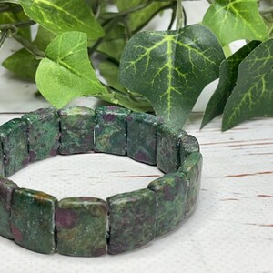 May include: A green and red stone bracelet. The bracelet is made of rectangular stones with a mottled pattern of green and red. The bracelet is displayed on a white surface with green leaves in the background.