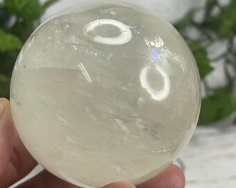 Op de afbeelding: Een heldere, bolvormige kristallen bol met interne adering en subtiele iriserende highlights. Het gepolijste oppervlak reflecteert licht, waardoor een zachte gloed ontstaat. De kristal wordt in een hand gehouden, wat de grootte en helderheid aantoont.