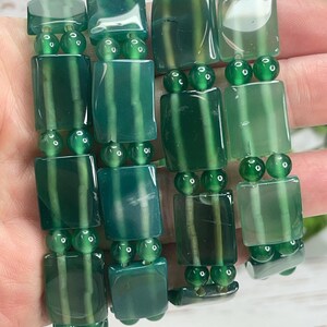 May include: A close-up of green gemstone bracelets. Each bracelet features rectangular and round beads in varying shades of green. The rectangular beads have a striped pattern, and the round beads are smaller and more uniform in color. The bracelets are held in a hand.