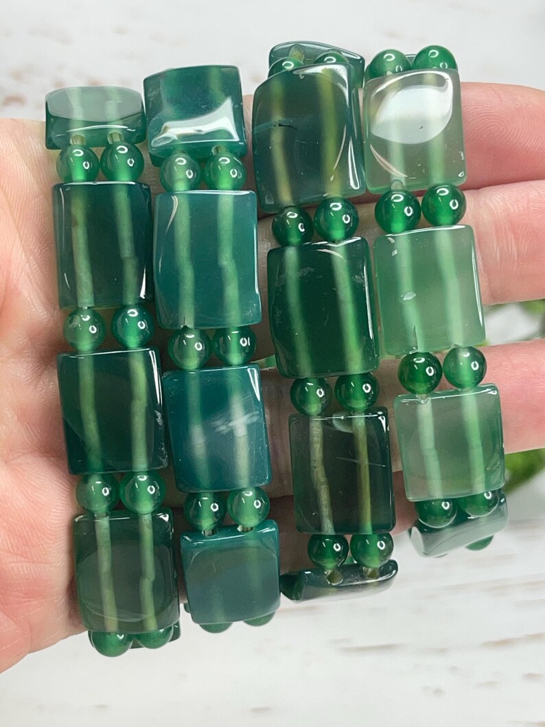 May include: A close-up of green gemstone bracelets. Each bracelet features rectangular and round beads in varying shades of green. The rectangular beads have a striped pattern, and the round beads are smaller and more uniform in color. The bracelets are held in a hand.