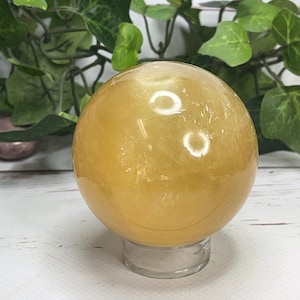 May include: A golden yellow crystal sphere, resting on a clear glass stand. The polished surface of the sphere reflects light, creating highlights. Green leafy vines are in the background, adding a natural element. This decorative sphere is a unique home decor item.