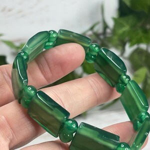 May include: A green gemstone bracelet featuring rectangular and round beads. The bracelet is made of polished, translucent green stones, strung together with small, round connecting beads. The bracelet is shown against a soft, blurred background.