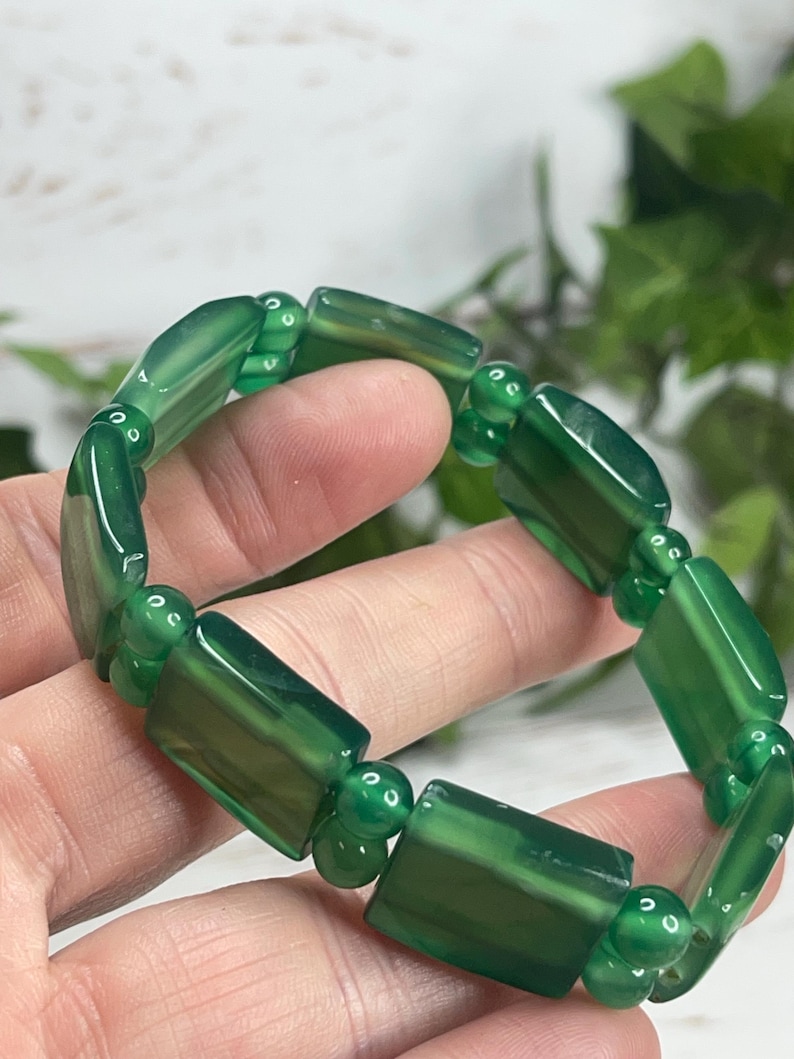 May include: A green gemstone bracelet featuring rectangular and round beads. The bracelet is made of polished, translucent green stones, strung together with small, round connecting beads. The bracelet is shown against a soft, blurred background.