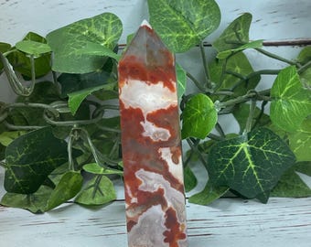 May include: A red and white agate crystal point, also known as a wand or obelisk. The crystal is polished and has a smooth, glassy finish. It is a natural stone with unique patterns and colors.