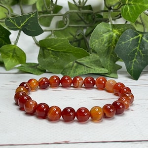 May include: A beaded bracelet composed of round, polished carnelian stones in shades of red, orange, and amber. The bracelet is strung on an elastic band and rests on a white wooden surface, with green foliage in the background.
