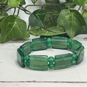 May include: A green beaded bracelet with rectangular and round beads. The bracelet is made of polished, translucent green stones. The background includes green leaves and a white wooden surface.