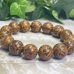 May include: A beaded bracelet featuring spherical beads with a brown and yellow patterned design. The bracelet is displayed on a light-colored surface with green foliage in the background. The beads are approximately 0.75 inches in diameter.