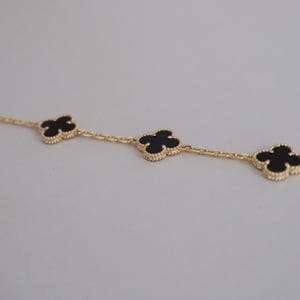 May include: A gold chain bracelet featuring three evenly spaced four-leaf clover motifs. Each clover motif is outlined with a beaded gold edge and contains a smooth, flat black inlay. The chain consists of small, polished gold oval links.