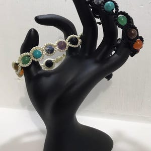 May include: Three bracelets featuring colourful gemstone beads and black macrame cord. The bracelets are displayed on a black hand-shaped stand.