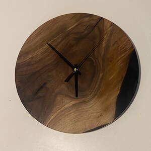 Puede incluir: Un reloj de madera redondo con agujas negras y un centro dorado. La esfera del reloj est&aacute; hecha de madera oscura con un patr&oacute;n de veta natural. El reloj est&aacute; montado en una pared blanca.