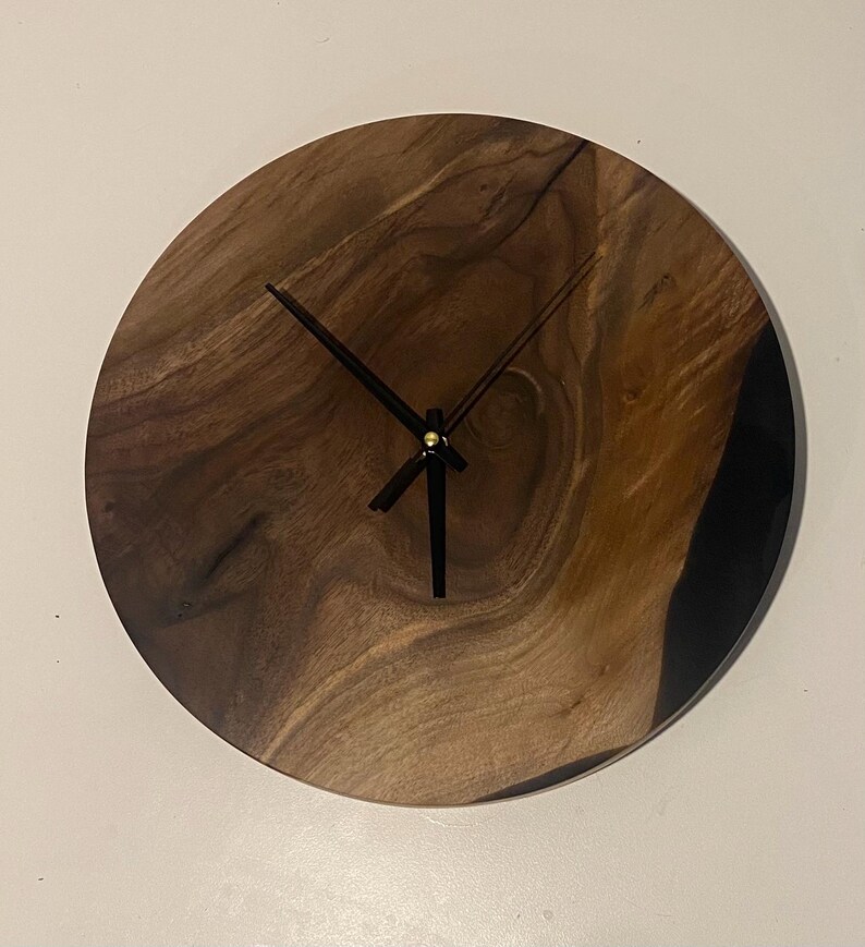 Puede incluir: Un reloj de madera redondo con agujas negras y un centro dorado. La esfera del reloj est&aacute; hecha de madera oscura con un patr&oacute;n de veta natural. El reloj est&aacute; montado en una pared blanca.