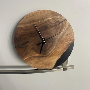 Puede incluir: Un reloj redondo de madera con agujas negras y un borde de resina epoxi negra. El reloj est&aacute; montado en una pared blanca.