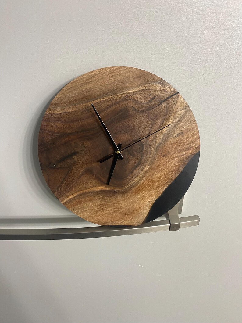 Puede incluir: Un reloj redondo de madera con agujas negras y un borde de resina epoxi negra. El reloj est&aacute; montado en una pared blanca.