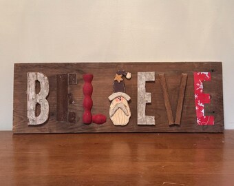 Puede incluir: Letrero de madera con la palabra "BELIEVE". Las letras son de madera, pintadas de rojo y con tela estampada. La "I" es un elemento decorativo de madera roja. La "E" presenta una figura de Papá Noel. Decoración navideña.
