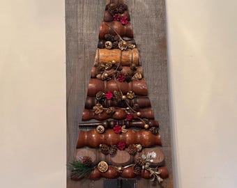Puede incluir: Un adorno de pared rústico con forma de árbol de Navidad hecho de madera, piñas y elementos decorativos. El árbol es marrón y beige, con una estrella en la parte superior. El fondo es una tabla de madera desgastada. La letra "C" está en la esquina inferior derecha.