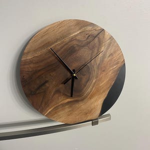 Puede incluir: Un reloj de pared redondo de madera con manecillas negras y un borde de resina epoxi negra. El reloj est&aacute; montado en un soporte de metal plateado.