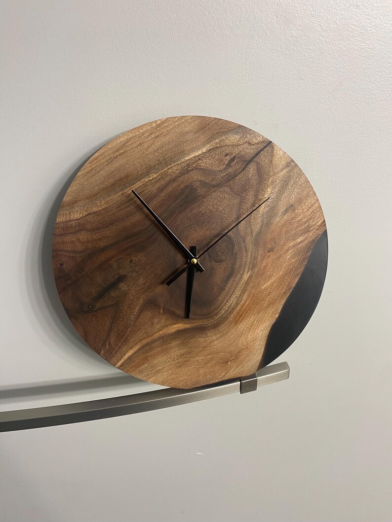 Puede incluir: Un reloj de pared redondo de madera con manecillas negras y un borde de resina epoxi negra. El reloj est&aacute; montado en un soporte de metal plateado.