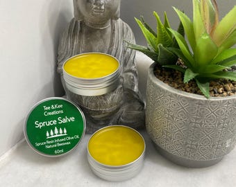 Puede incluir: Dos pequeños botes redondos de bálsamo amarillo, etiquetados con "Spruce Salve" y el texto "Spruce Resin Infused Olive Oil, Natural Beeswax, 60ml".