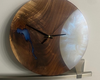 Puede incluir: Un reloj redondo de madera con una incrustación de resina blanca. La esfera del reloj tiene agujas negras y un centro dorado. La madera es de color marrón oscuro con una incrustación de resina azul.