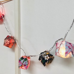 Può includere: Una ghirlanda di luci con lanterne di carta. Le lanterne sono fatte di tessuto con motivi floreali in tonalità rosa, blu e verde. Le luci sono infilate su un filo d'argento.