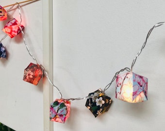 Puede incluir: Una cadena de luces con farolillos de papel. Los farolillos están hechos de tela con estampados florales en tonos rosa, azul y verde. Las luces están enhebradas en un alambre plateado.