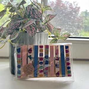 Puede incluir: Un juego de diez pinzas de madera para la ropa empaquetadas en una funda de plástico transparente, con varios patrones coloridos que incluyen globos, regalos y rayas rosas y azules, con una pequeña pegatina redonda que dice thank you for supporting my small business.