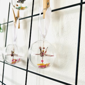 May include: Clear glass baubles with origami cranes, star-shaped confetti, and white ribbon hangers. The baubles are clipped to a black wire grid with wooden pegs. The cranes are pink, red, and gold.