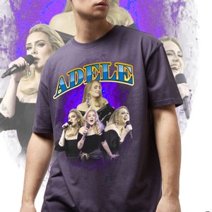 K&ouml;nnte beinhalten: Ein dunkelviolettes T-Shirt mit dem Namen "ADELE" in einem gold-blauen Bogen-Design. Das Shirt zeigt mehrere Bilder von Adele beim Singen, vor einem lila-wei&szlig;en Hintergrund. Das Design hat eine Retro-&Auml;sthetik.