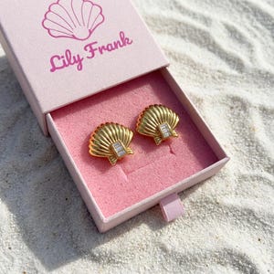 May include: A pair of gold-colored seashell-shaped earrings with rectangular, clear stone accents, presented in a pink box. The box is labeled "Lily Frank" with a seashell graphic. The earrings are displayed on a bed of pink velvet.