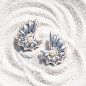 May include: A pair of silver-toned earrings shaped like stylized seashells, each featuring a single small white pearl set in the center. The metal surface has a polished, reflective finish with radiating ridges that mimic the natural texture of a shell.