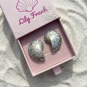 May include: Two iridescent, spiral-shaped shell earrings with a pearlescent finish and dark, oval-patterned markings. They are presented inside a light pink sliding jewelry box featuring a shell icon and the text Lily Frank printed in dark pink.