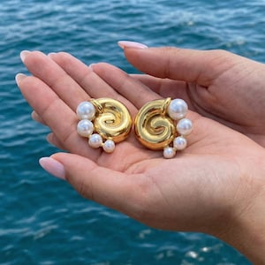 May include: A pair of gold-coloured, spiral-shaped earrings with a cluster of white pearls. The earrings are held in an open hand against a backdrop of blue water. The earrings have a polished, reflective surface.