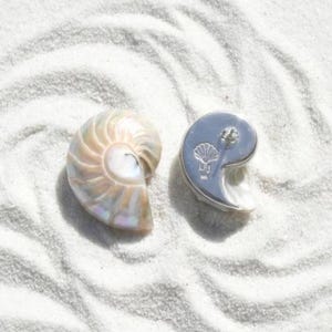 May include: A nautilus shell and a silver-colored earring in the shape of a yin-yang symbol with a seashell design, resting on white sand. The earring has a post for pierced ears.