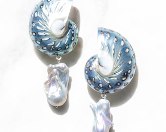 Può includere: Un paio di orecchini fatti a mano con un design a conchiglia blu e bianco e una perla pendente di forma irregolare. Gli orecchini hanno un attacco in metallo color argento. Il design a conchiglia ha un motivo maculato.