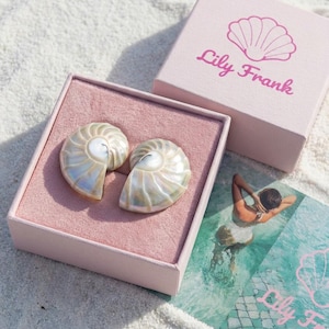 May include: Two nautilus shell earrings displayed in a pink box with a matching lid. The box is on a sandy surface. The brand name "Lily Frank" is printed on the box and a card.
